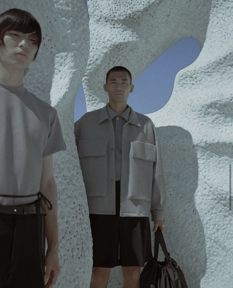 Two Asian men pose in a fashion shot outdoors. One holds a bag, standing amidst a textured, porous sculpture against a blue sky.
