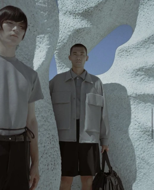 Two Asian men pose in a fashion shot outdoors. One holds a bag, standing amidst a textured, porous sculpture against a blue sky.