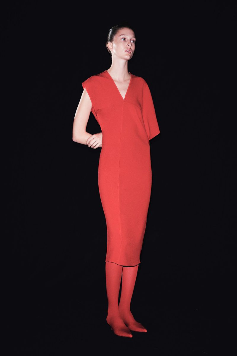 A full shot of a young adult woman posing in a red, v-neck dress with red stockings against a solid black backdrop in a studio.