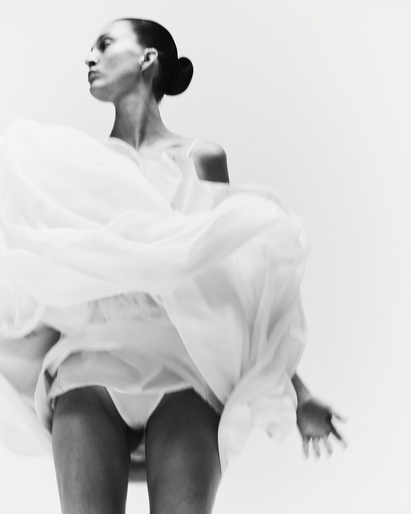Low-angle shot of a woman with slicked-back hair in a bun dancing in a voluminous white dress against a white studio backdrop.