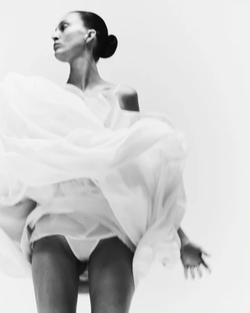 Low-angle shot of a woman with slicked-back hair in a bun dancing in a voluminous white dress against a white studio backdrop.