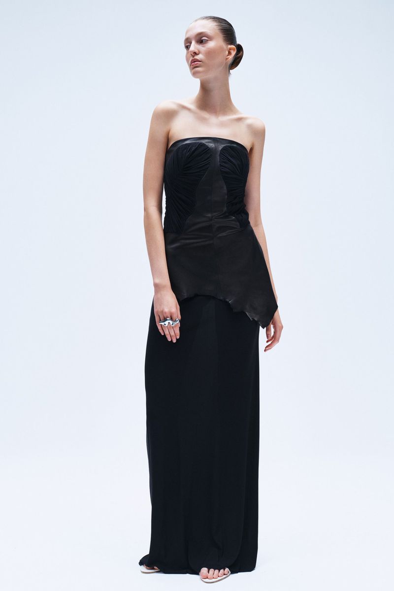 Full shot of a slender woman posing in a strapless black gown with a leather bodice against a bright white backdrop in a fashion setting.