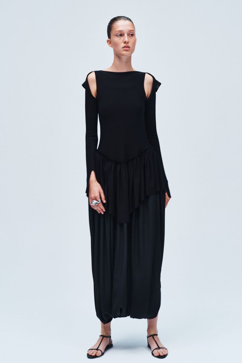 A full-length portrait of a woman in a black, avant-garde dress with long sleeves and t-strap sandals against a bright, neutral background.