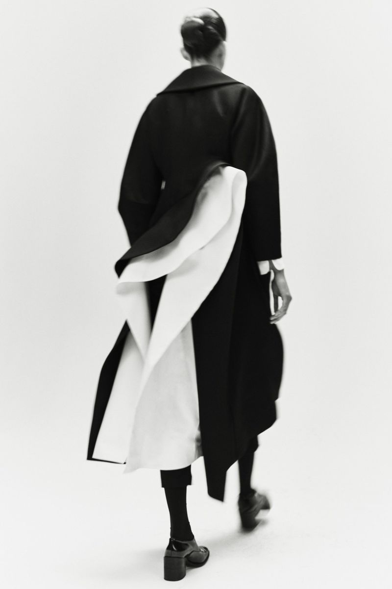 Fashionable adult female model walks away wearing a black and white coat, hosiery, and high heels in a light studio.