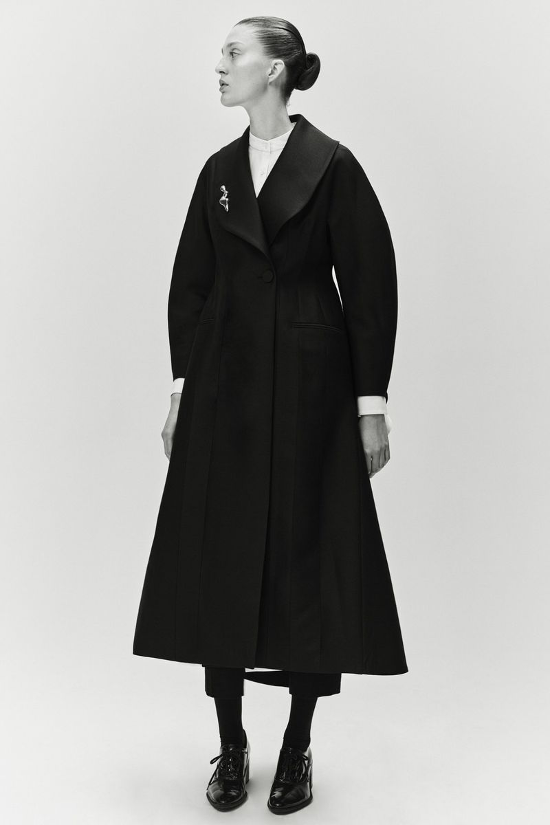 A single female model showcases a long black coat with wide lapels, styled with dark tights and shoes, against a plain white backdrop.