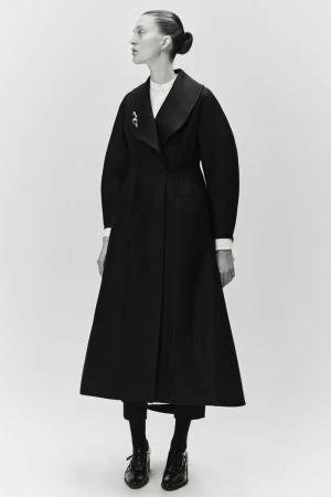 A single female model showcases a long black coat with wide lapels, styled with dark tights and shoes, against a plain white backdrop.