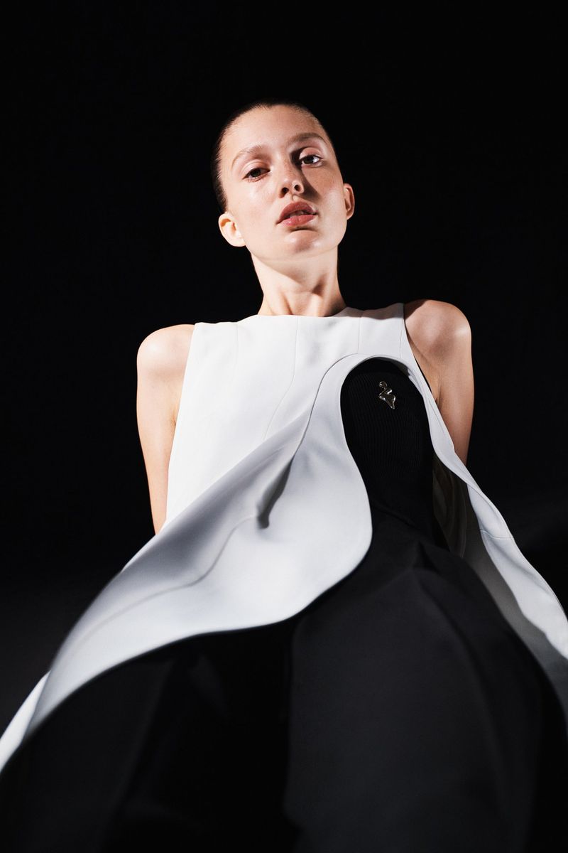 Woman in sleeveless white and black dress stands in front of black background in a fashion style portrait with eye-level angle.