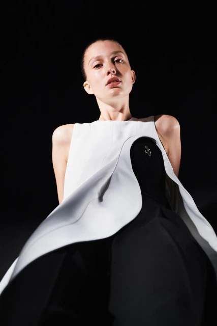 Woman in sleeveless white and black dress stands in front of black background in a fashion style portrait with eye-level angle.