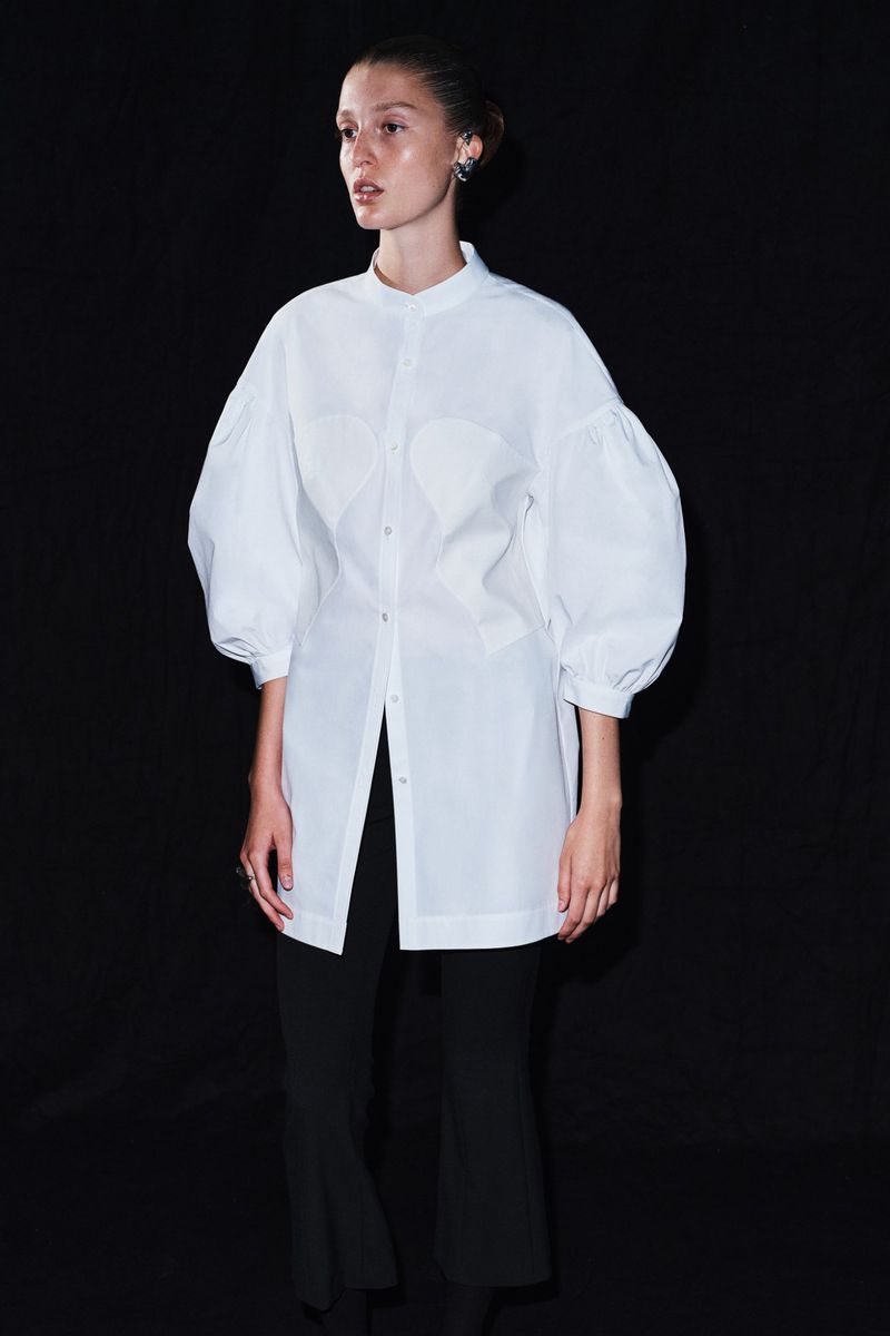 A woman stands in a photo studio wearing a long white button-down shirt with puffy sleeves, black pants, and jewelry against a black background.