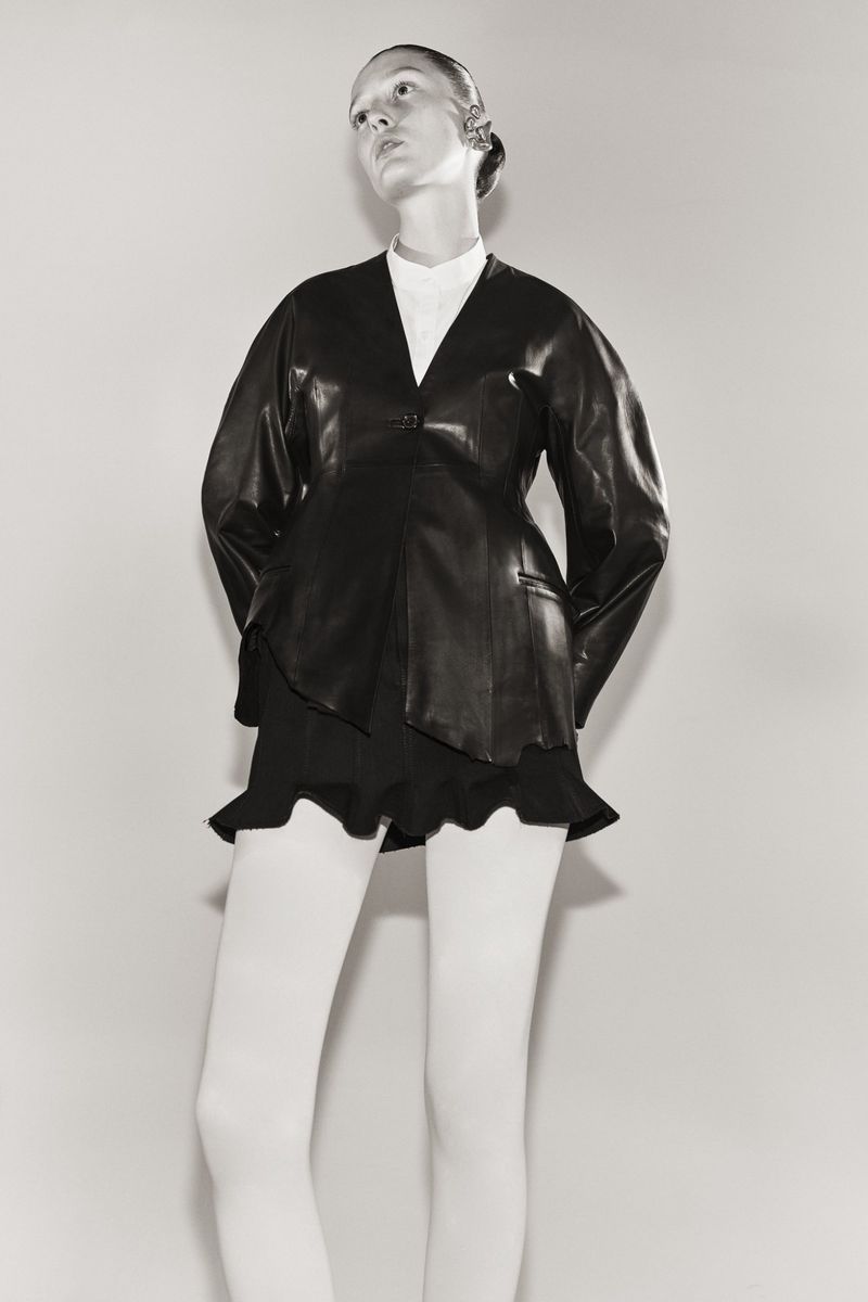 Fashionable adult female model in a studio setting, wearing a black leather jacket, a short skirt, and white tights against a grey backdrop.