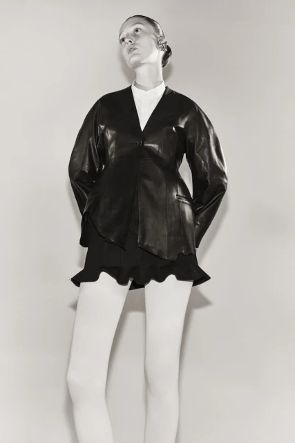 Fashionable adult female model in a studio setting, wearing a black leather jacket, a short skirt, and white tights against a grey backdrop.
