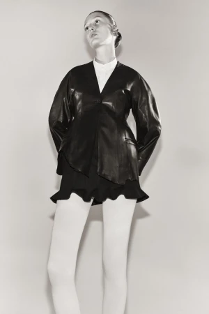 Fashionable adult female model in a studio setting, wearing a black leather jacket, a short skirt, and white tights against a grey backdrop.