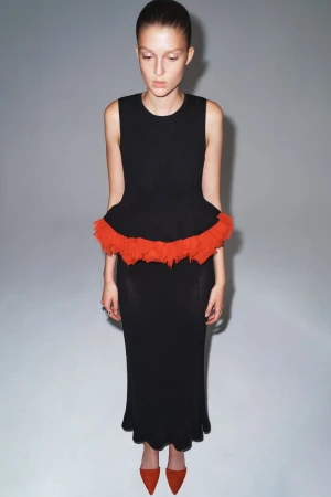 Model wearing a sleeveless black dress with an orange ruffled peplum hem against a grey studio backdrop.