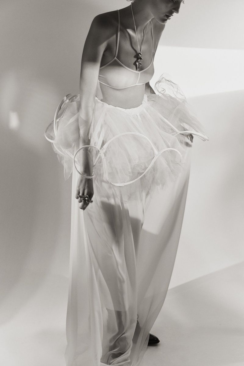 Monochrome portrait of a slender woman in a bra top, tulle skirt, and long sheer skirt, against a simple white backdrop in a studio setting.