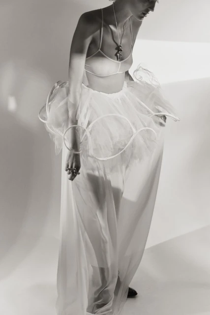 Monochrome portrait of a slender woman in a bra top, tulle skirt, and long sheer skirt, against a simple white backdrop in a studio setting.