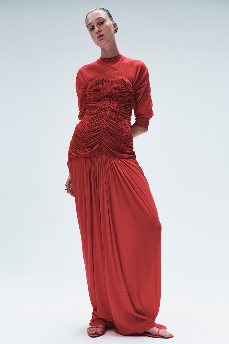A slender woman poses in a red floor-length dress with a shirred bodice against a gradient white backdrop in a fashion editorial shot.