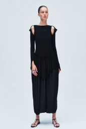 Model standing against a light grey background wearing a long-sleeved black dress with cutout shoulders and drop-waist ruffled skirt.