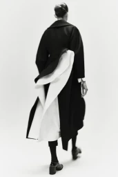 Model viewed from behind walking away in an architectural black coat with cascading white drapes against a white backdrop.
