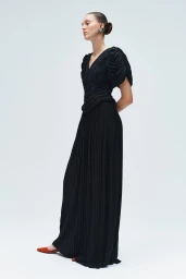 Model wearing a textured black short-sleeve top and floor-length pleated maxi skirt with rust shoes against a white studio backdrop.