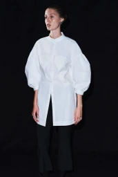 Model Stinne Trappeniers wearing a white puff-sleeve button-down tunic and black trousers against a solid black backdrop.