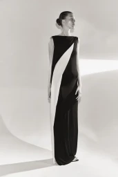 Model wearing a long black sleeveless gown with a diagonal white drape panel against a white studio background.