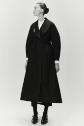 Model posing in a structured black A-line coat and oxford shoes against a seamless studio backdrop for Ashlyn S/S25.