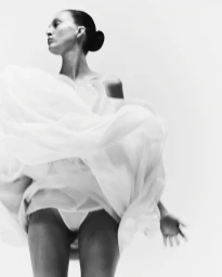 Low-angle shot of a woman with slicked-back hair in a bun dancing in a voluminous white dress against a white studio backdrop.