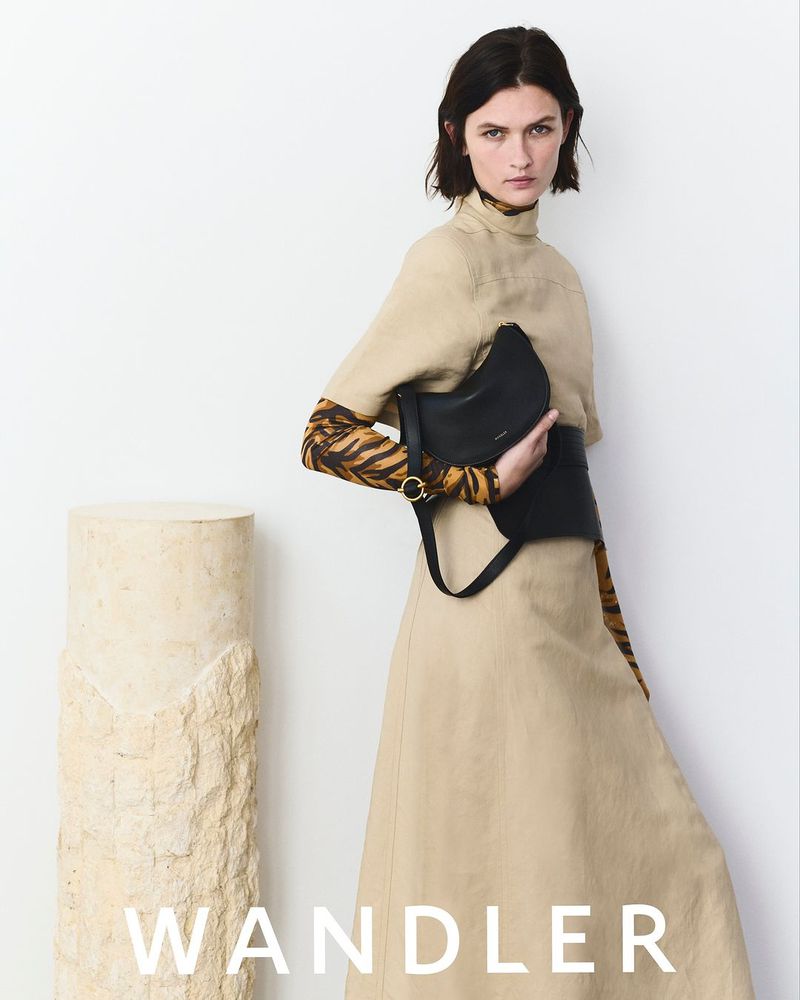 Fashion shot of a female model posing in a beige dress with a black handbag, next to a stone column against a white background.