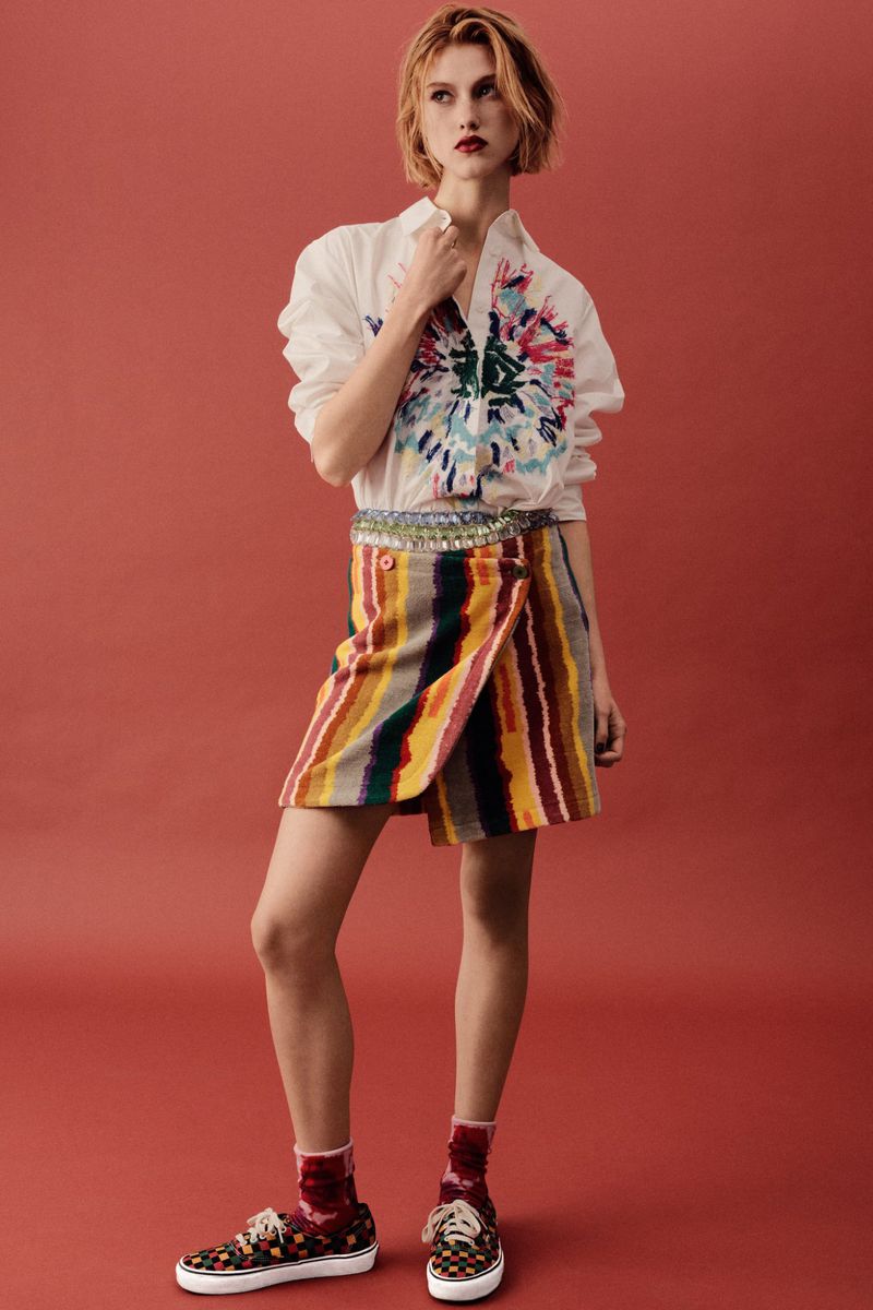 Model Gracie Rae wearing a white printed shirt and colorful striped wrap skirt against a terracotta studio background.