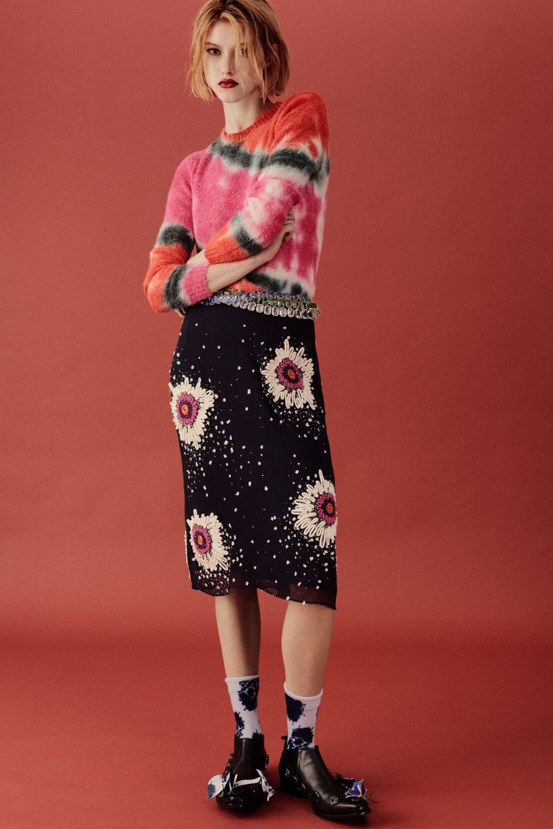 Full-length shot of a woman in a colorful tie-dye sweater and floral-print skirt standing in front of a solid coral background.
