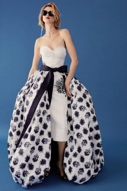 Woman wearing a strapless white dress with a full patterned skirt and black belt, sunglasses, and long earrings, posing against a blue background.