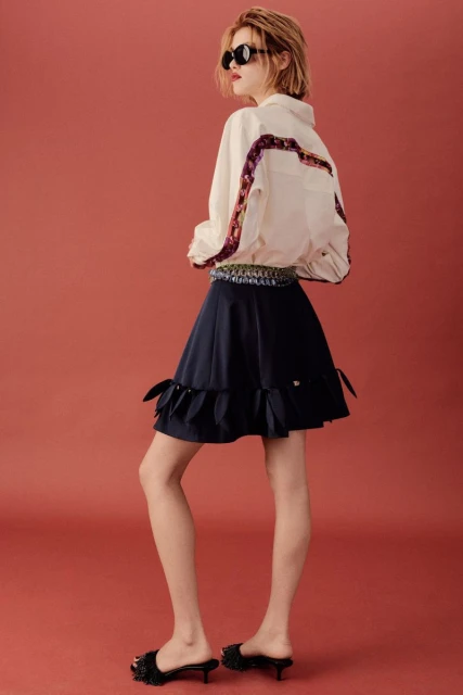 Young woman in stylish cream shirt and navy skirt poses in studio against a red background, wearing sunglasses and black heeled shoes.
