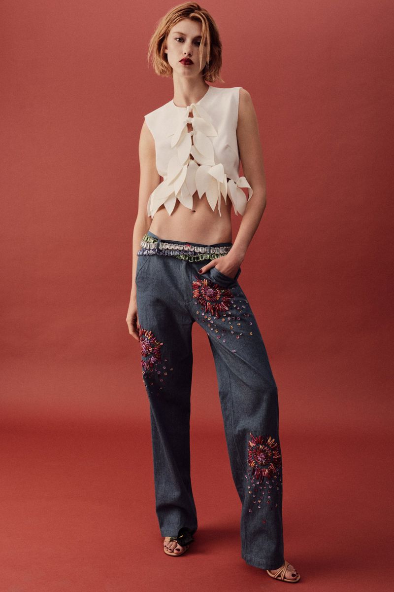 A young woman in a white vest with leaf details and blue embellished jeans strikes a fashion pose in a full shot against a red background.