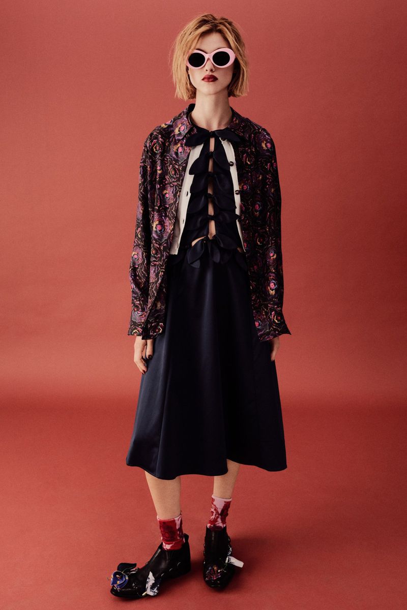 Young woman in a patterned jacket, dark dress, sunglasses, and statement shoes poses against a terra cotta background.