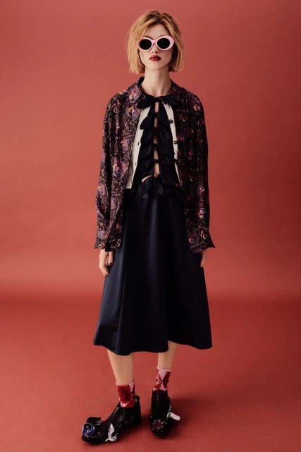 Young woman in a patterned jacket, dark dress, sunglasses, and statement shoes poses against a terra cotta background.