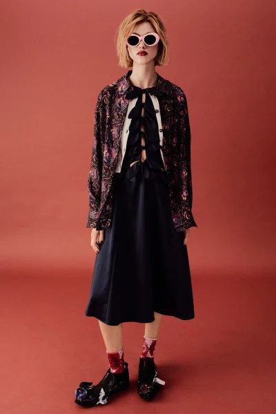 Model wearing patterned jacket over bow-detailed black dress with pink sunglasses and patterned socks against a terracotta seamless background.