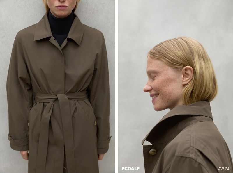 Woman wears a brown trench coat indoors against a neutral wall. The model is fair-skinned with freckles and blonde hair.