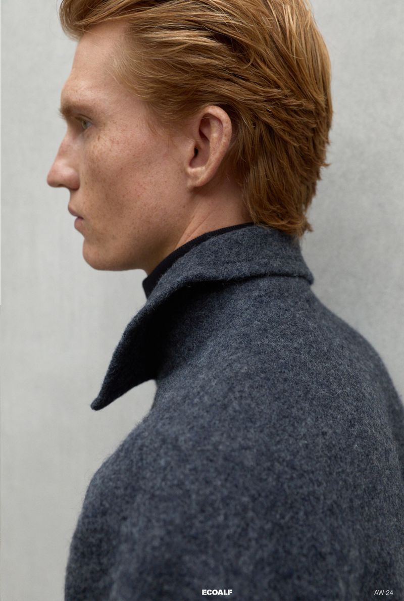 Profile shot of a fair-skinned adult male with red hair wearing a dark grey wool coat against a neutral grey background in a studio setting.