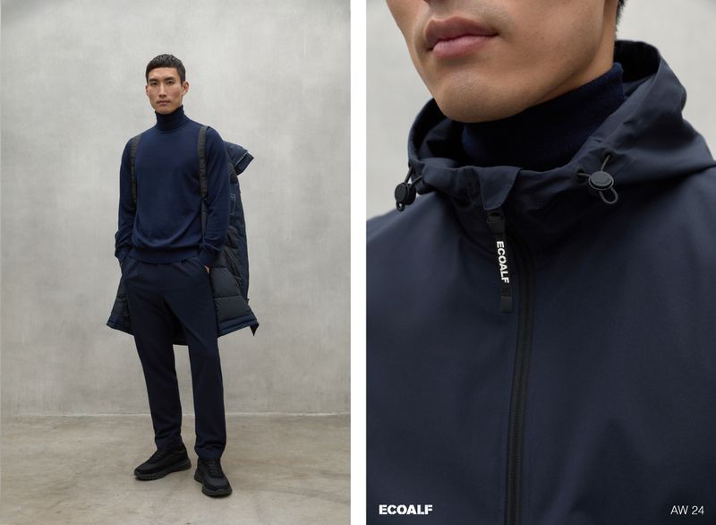 Man in a navy turtleneck, slacks, and shoes stands near a gray wall; a navy puffer coat hangs from his shoulder in the studio shot.