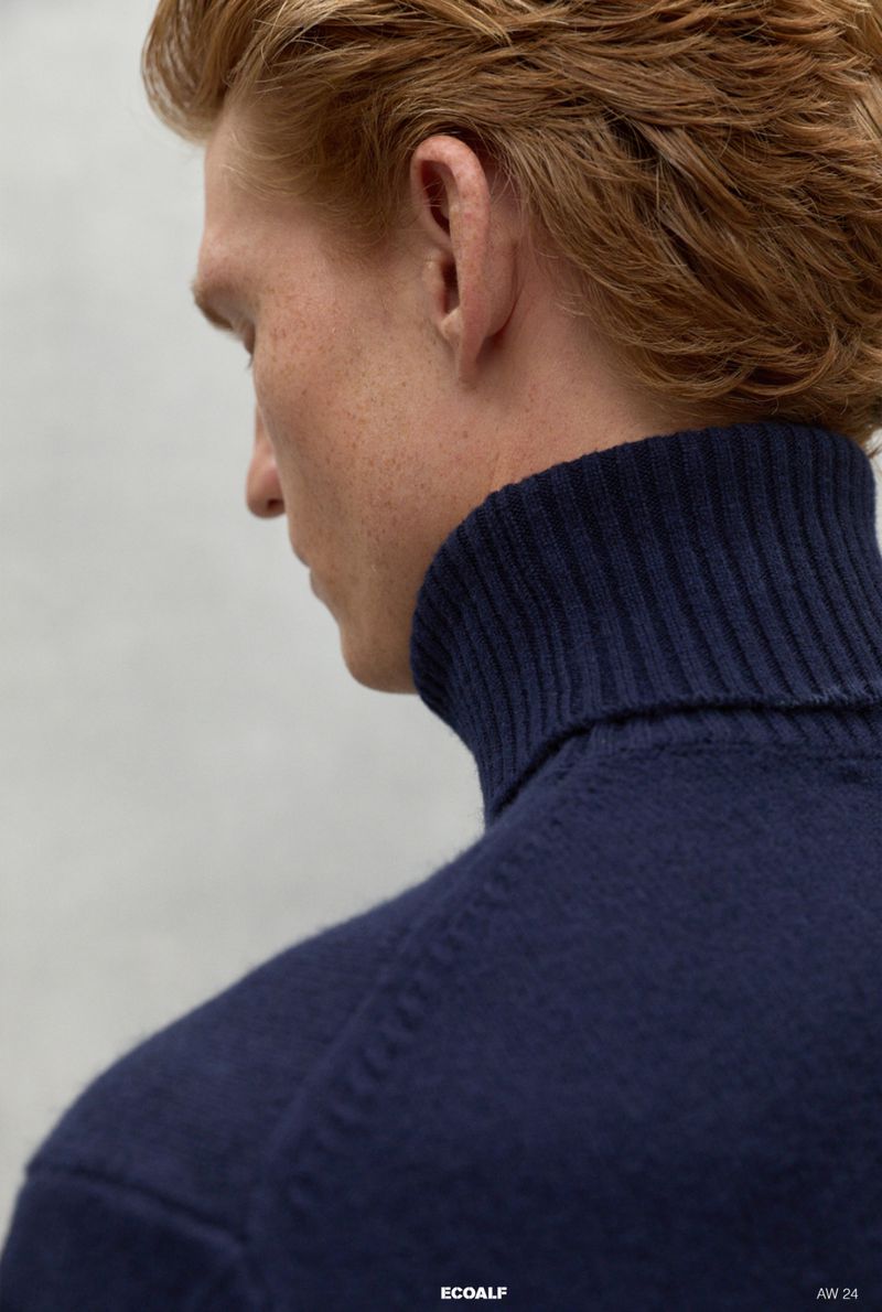 Close-up studio shot of a light-skinned adult male with red hair, wearing a navy blue turtleneck sweater against a neutral background.