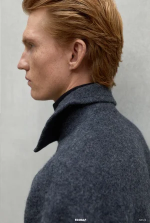 Profile portrait of a man with reddish-blonde hair wearing a textured charcoal wool coat against a pale grey backdrop.