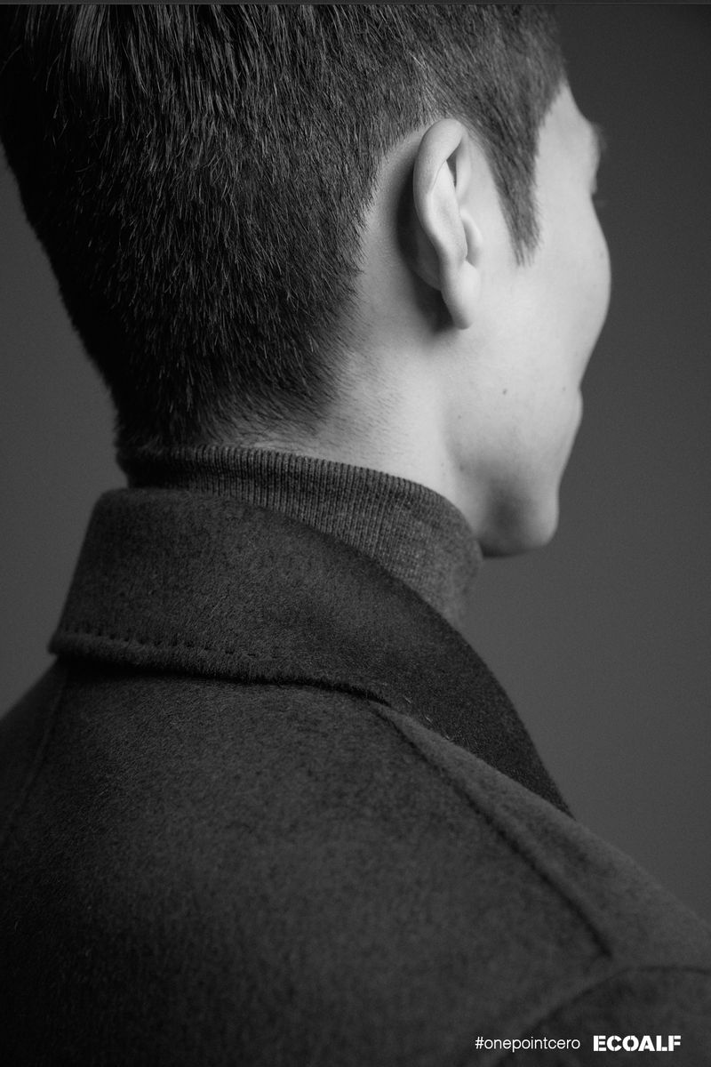 Monochrome portrait of an adult male wearing a wool coat and turtleneck sweater, posing in a photo studio with gray background.