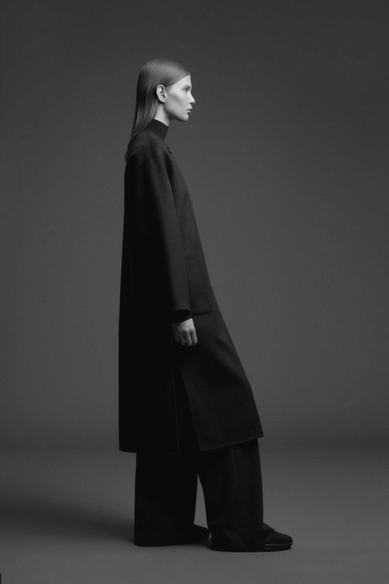 Model in profile wearing a structured full-length black coat and wide-leg trousers against a seamless dark grey studio backdrop.