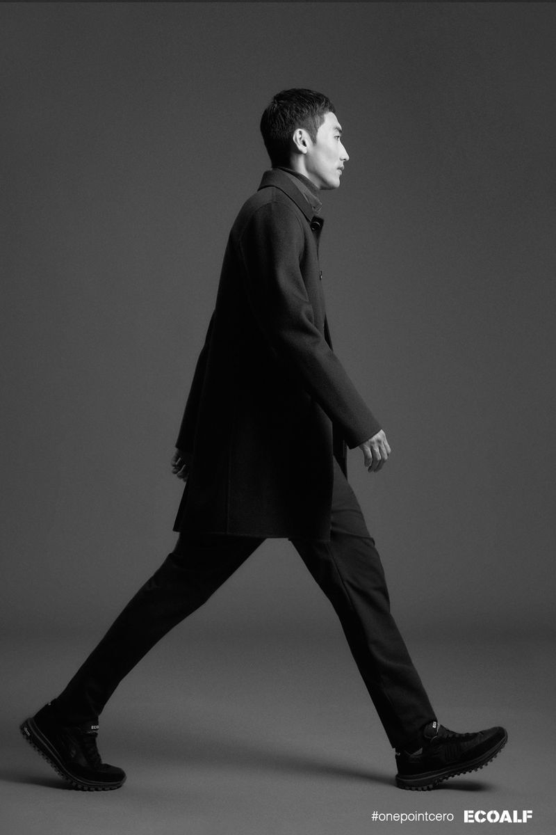 Monochrome fashion shot: Man in a dark coat and pants walks in profile view against a grey backdrop, showcasing a minimalist aesthetic.