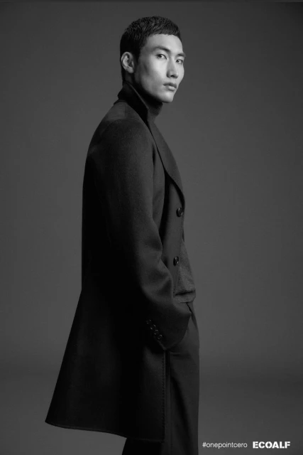 Portrait of a stylish Asian man in a long, dark overcoat and turtleneck, posing in a studio with a gray background, fashion photography.