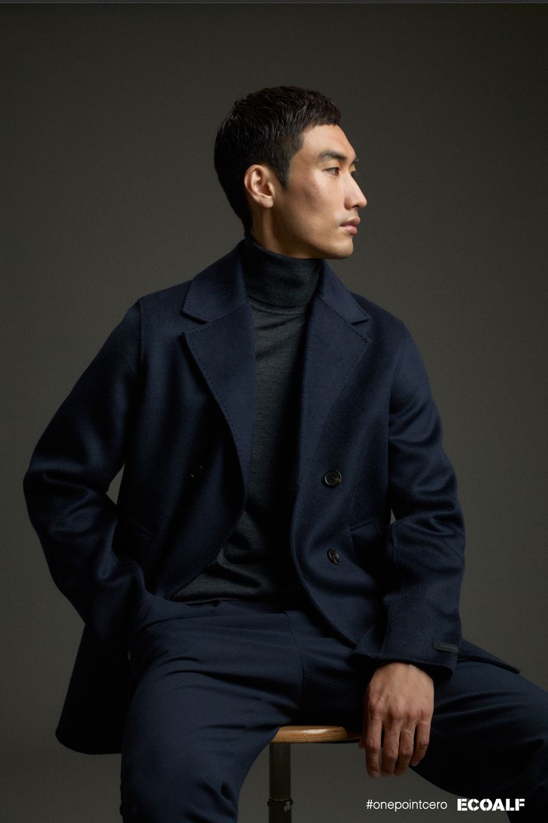 Portrait of an Asian adult male in a navy coat and trousers, sitting on a wooden stool in a photo studio against a grey backdrop.