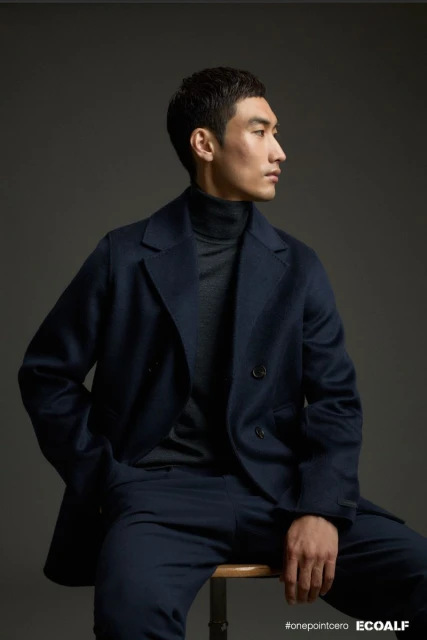 Portrait of an Asian adult male in a navy coat and trousers, sitting on a wooden stool in a photo studio against a grey backdrop.