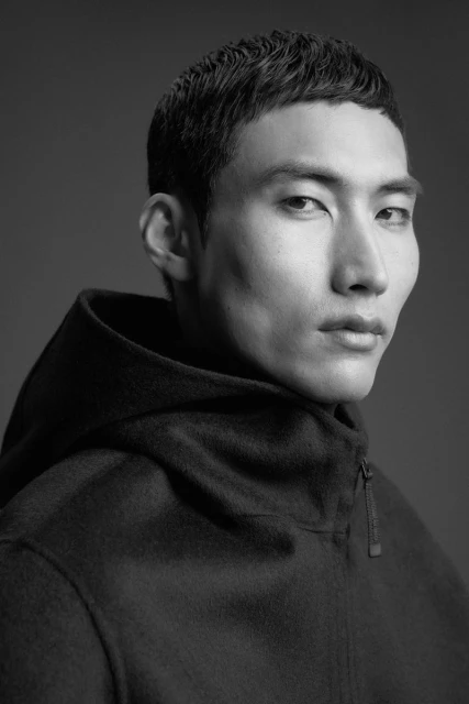 Monochromatic portrait of an Asian adult male in a hooded coat, looking directly at the camera with neutral expression against a grey backdrop.