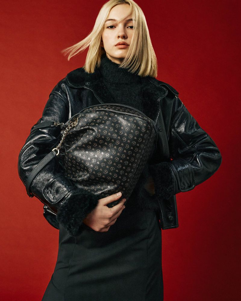 Young woman wearing a black leather jacket holds a patterned satchel bag against a red background in a fashion portrait.