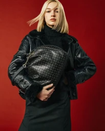 Blonde woman wearing a black leather jacket holding a monogrammed black handbag against a red background
